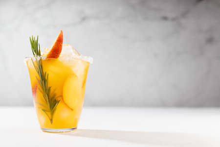 Homemade vitamin summer fresh orange peach lemonade with ice, green rosemary twig, sugar rim, fruit slices in misted glass on white wood table, soft light interior.の写真素材