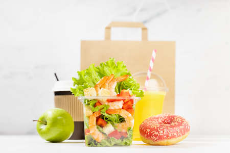 Healthy daily diet plan - set of orange juice, tropical salad with shrimps in plastic box, pink donut, packet in white interior. Advertising for restaurant take away, delivery food service.の写真素材