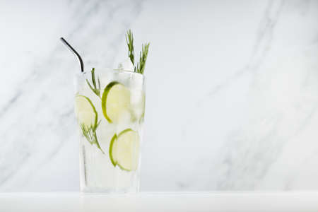 Fresh citrus infused homemade lemonade with ice cubes, lime slices, green rosemary twig, silver straw in soft light white kitchen interior in sunny day.の写真素材