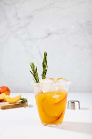 Cooking of fresh cold peach drink in glass with ice cubes, green rosemary, fruit slices, ingredients in soft light white kitchen interior, vertical.の写真素材