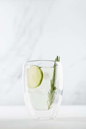 Detox cold lime lemonade with ice cubes, lime slices, rosemary twig in transparent mug on white wood table in sunlight, summer cocktail, vertical.の写真素材