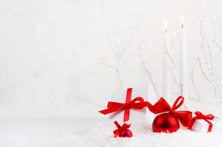 Christmas background with white burning candles, gift boxes, red decorations - glossy balls with satin ribbons in decorative soft light white winter forest with frosty branch and snow, copy space.の写真素材