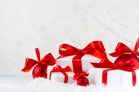 Christmas festive background in white and red color - shimmer balls and gift boxes with satin ribbon closeup in snowdrift under frosty white branches, snow in winter forest, copy space.の写真素材