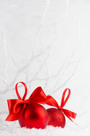Christmas decoration - two rich red glossy glitter balls with satin ribbon in decorative soft light white winter forest with frosty branch, snow, copy space, vertical. Festive New Year background.の写真素材