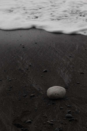 Wet black volcanic sand on seashore with white foam of wave as line and white stone, zen, texture, background, vertical.の写真素材