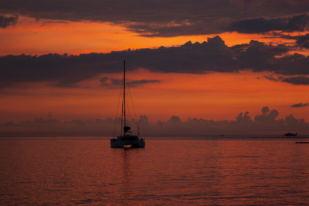 Majestic saturated sea landscape - colorful sunset in ocean with golden sky, dark clouds, reflections in calm water, orange sunbeams, silhouette floated yacht. Marine landscape on indonesian island.の写真素材