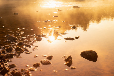 Warmy golden sunlight on quiet water surface of lake, orange haze, ripple sun glares, blinks, reflection, stones on shore in early morning, idyllic, tranquil wild nature background, texture, detail.の写真素材