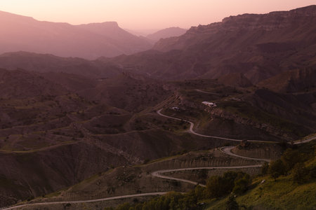 Idyllic landscape  - silhouette of high mountain green ridges in pink pastel sunrise sunlight, mist, serpentine road, village away in early summer morning, panorama view. Majestic travel in Dagestan.の写真素材