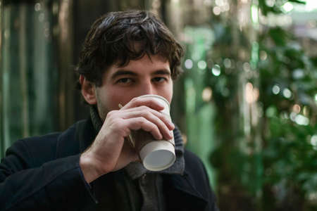Young handsome man in coat drinking coffee looking at cameraの写真素材
