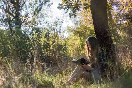Girl sitting under a tree reading book in summer forestの写真素材