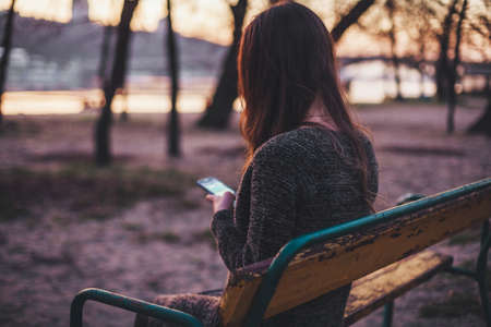 Young woman sitting on the bench near the river typing a message on smart phoneの写真素材