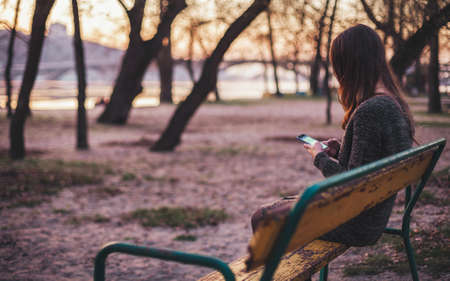 Young woman sitting on the bench near the river typing a message on smart phoneの写真素材