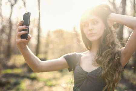 Happy girl making selfie photo on smartphone in the park. Focus on phoneの写真素材