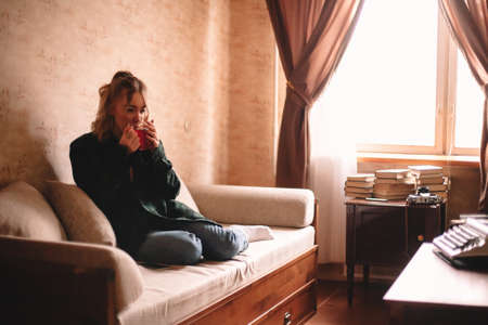 Young thoughtful woman drinking tea while sitting on sofa in living room at homeの写真素材