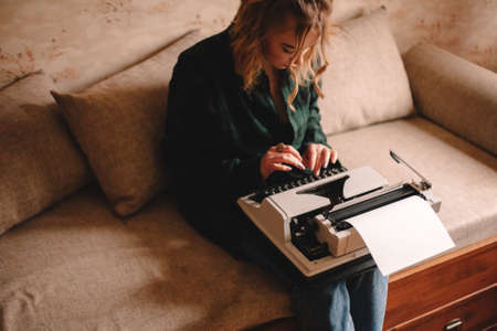 Young female writer using typewriter while sitting on sofa at homeの写真素材