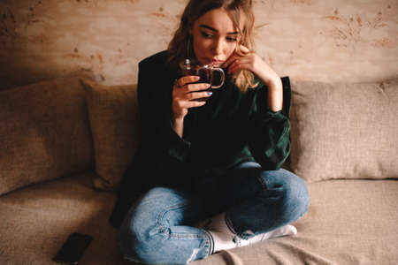 Young thoughtful woman drinking tea while sitting on sofa at homeの写真素材