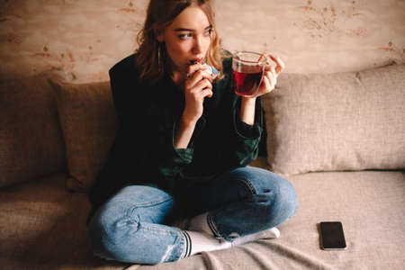 Young thoughtful woman drinking tea and eating chocolate while sitting on sofa at homeの写真素材