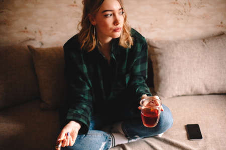 Young thoughtful woman holding cup of tea and chocolate while sitting on sofa at homeの写真素材