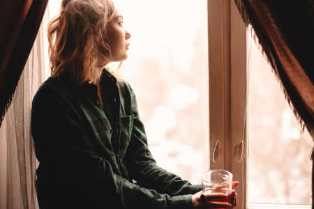 Young thoughtful woman drinking tea and looking through window while sitting on windowsill at home