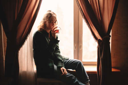 Young thoughtful woman drinking tea and looking through window while sitting on windowsill at homeの写真素材