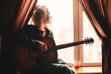 Young woman looking through window while sitting on windowsill with guitar at homeの写真素材