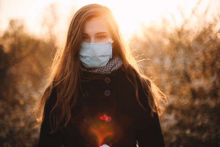 Portrait of sick young woman wearing protective face medical mask standing in park at sunset during springの写真素材