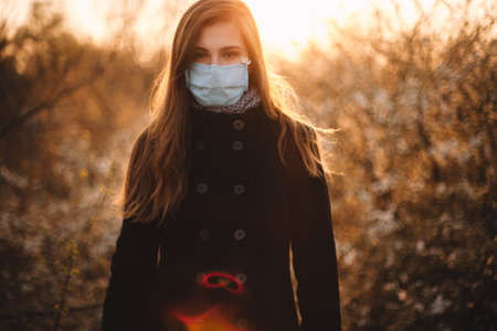 Portrait of sad young woman wearing protective face medical mask standing in park at sunset during springの写真素材