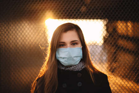 Portrait of young woman wearing protective face medical mask while standing against fence at sunsetの写真素材