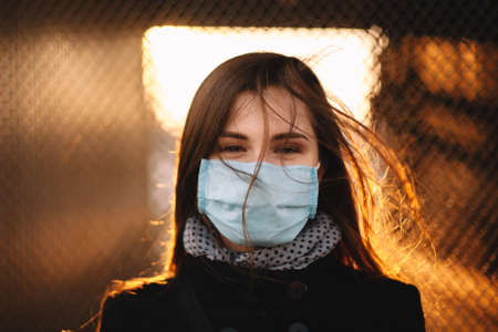 Portrait of young woman wearing protective face medical mask while standing against fence at sunsetの写真素材