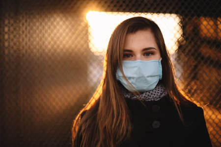 Portrait of young woman wearing protective face medical mask while standing against fence at sunsetの写真素材