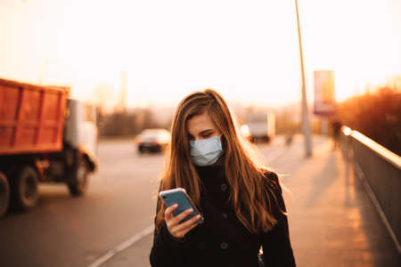 Happy young woman wearing protective face medical mask using smart phone while walking on empty sidewalk on bridge in city at sunsetの写真素材