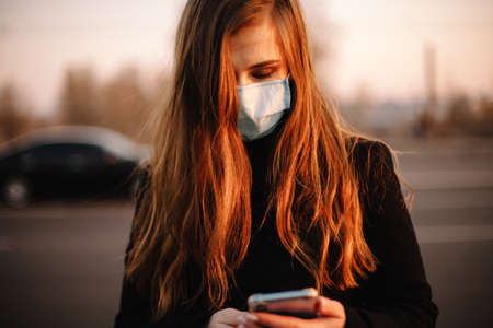 Portrait of young woman wearing protective face medical mask using smart phone while standing on street in city at sunsetの写真素材