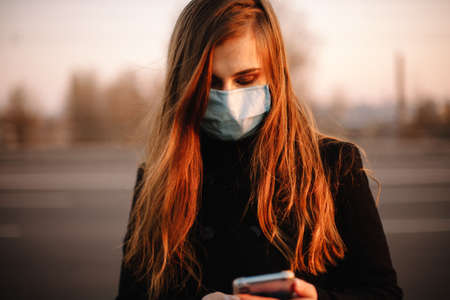 Portrait of young woman wearing protective face medical mask using smart phone while standing on street in city at sunsetの写真素材