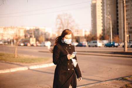 Young woman wearing protective face medical mask while using smart phone walking on street in cityの写真素材