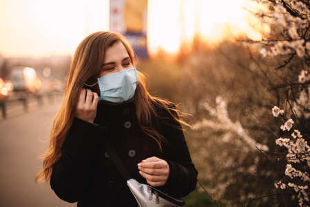 Happy young woman putting on face medical mask while standing on street in city during sunset in springの写真素材