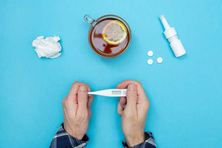 Man is holding a thermometer. Flu season. Nearby are pills, nasal spray, used wipes, and lemon tea. Catch a cold. Disease. Flat lay. Seasonal cold concept.の写真素材