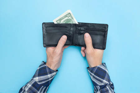 Young man holds a wallet in which there are only two dollars. Blue background. Bankrupt. Money is tight. Importance of savings. Financial aid concept.の写真素材