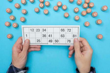 Man holds a card for lotto or bingo and there are many wooden barrels with numbers around on a blue...