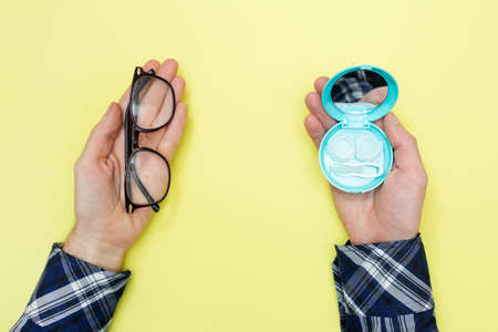 Man holds glasses for vision and a set of contact lenses for the purpose of choosing what to use. yellow background. flat layの写真素材