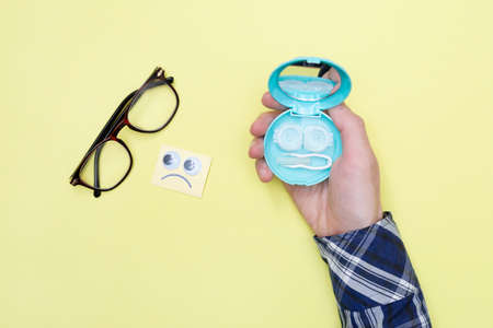 Sad emoticon looks at glasses and a man is holding a set for contact lenses on a yellow background....