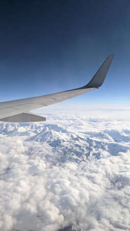 View from the window on the wing of the plane, blue sky, clouds and mountains in the snow. Place for...