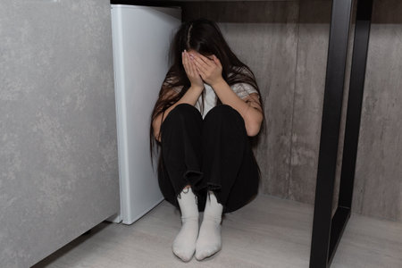 Frightened girl sits under the table covering her face with her hands. Stress after a domestic conflict.の写真素材