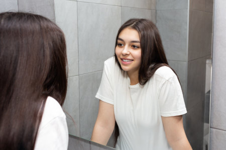 Smiling young girl looks in the mirror. Girl with long dark hair in the bathroom looks at herself in the mirror.の写真素材