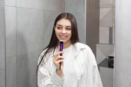 Beautiful brunette with long hair sings in the bathroom using a makeup brush. Concept of a good mood in a beautiful girl.の写真素材
