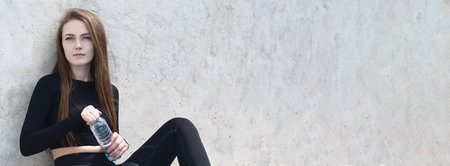 Young athletic woman holds a bottle of water to drink water during a break from training. Outdoor fitness training. Banner with space for text. Concrete background. Copy space.の写真素材