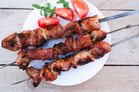 Juicy meat on skewers with tomatoes and herbs on a white plate on a wooden background. Kebab of meat is fried on the grill.の写真素材