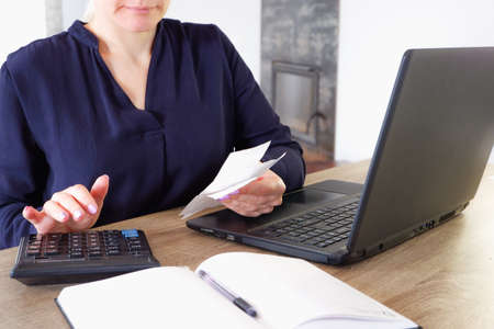 A young attractive woman, in a blue dress, sitting at a table with a laptop and a calculator, fills out tax reports. Calculation of payments. Annual tax return submission. Home environment, near the fireplace in the background. Remote work in quarantine.の写真素材