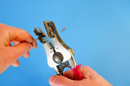The electrician removes the insulation from the wire using a special stripper tool. Close-up on hands on a blue background.の写真素材