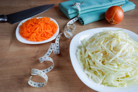 Healthy eating. Raw, chopped vegetables for salad on white plates. Cabbage, carrots, onions, measuring tape on a wooden tableの写真素材