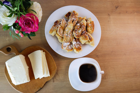 Home-baked brushwood during quarantine. Homemade dessert preparation. Homemade feta cheese, a cup of coffee on a wooden table top. Concept on the background of a white brick wall and a bouquet of roses.の写真素材